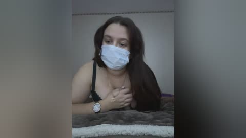 Media: Video of a woman with long brown hair, wearing a black bra, white mask, and wristwatch, lying on a gray bed, indoors.