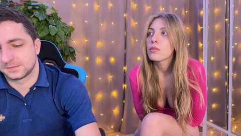 Media: A video of a man in a blue polo shirt and a woman in a pink top, both looking contemplative, in a cozy, softly lit room with string lights and a green plant.
