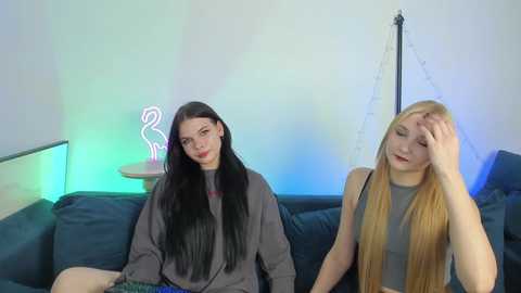 Video of two young women, one with long black hair, one with long blonde hair, sitting on a blue couch, in a modern living room with neon lights and fairy lights.