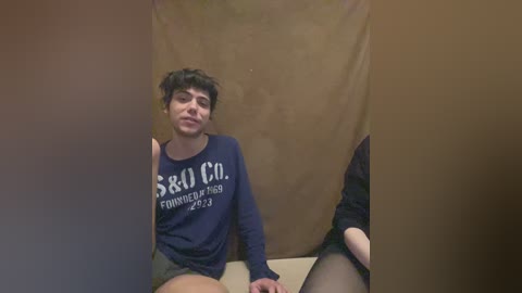 Video of a young person with short black hair, wearing a navy blue \"SARO CO\" t-shirt, gray shorts, and black stockings, sitting against a brown curtain backdrop.