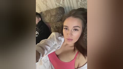 Media: Video of a young woman with long brown hair, light skin, and pink tank top, sitting on a patterned couch. A man in a black jacket is partially visible in the background.