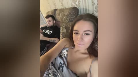 Media: Video of a woman in a gray patterned dress and a man in a black T-shirt with white letters, sitting on a brown sofa in a room with patterned wallpaper.