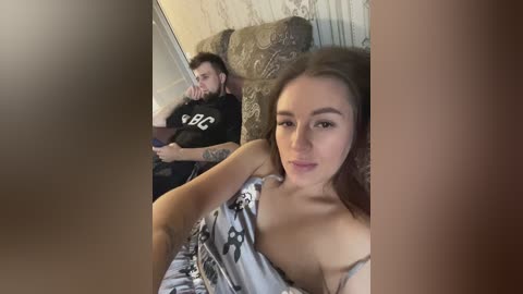 Media: Video of a young woman with light skin and straight brown hair, wearing a sleeveless floral dress, sitting on a couch, looking at a man with a beard in a black T-shirt and dark pants.