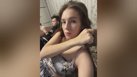 Media: A video of a young woman with long brown hair, wearing a floral top, taking a selfie in a dimly lit room with a man in the background.