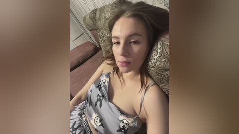 Media: A video of a young Caucasian woman with fair skin, medium brown hair, and light eyes, lying on a couch in a dimly lit room. She wears a gray, patterned camisole and matching pants.