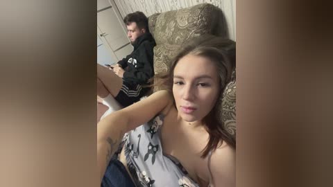 Media: Video of a young woman in a floral tank top, sitting on a patterned couch, looking towards a man in a hoodie, playing a video game in a dimly lit room.