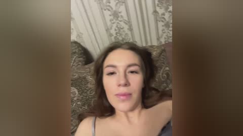 Media: Video of a young woman with light skin, brown hair, and subtle makeup, lying on a patterned bedspread, wearing a blue top. The background features a floral wallpaper.