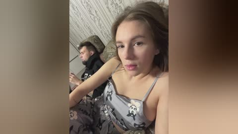 Media: A video shows a young woman with light skin and straight brown hair, wearing a light blue tank top with cartoon panda print, lying on a couch in a room with patterned wallpaper. Her boyfriend, with short brown hair, is next to her.