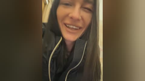 Media: A video of a Caucasian woman with long, straight, dark hair, wearing a black jacket, smiling broadly with eyes closed, standing in front of a blurred, beige wall.