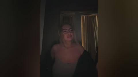 A video of a young woman with long blonde hair, wearing glasses, a black jacket, and a white top, standing in a dimly lit hallway.