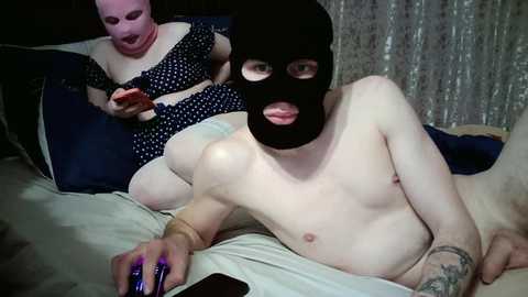A video of a shirtless man with a black mask and purple nails lying on a bed, while a woman in a polka dot dress sits next to him, both appearing to be in a domestic setting.