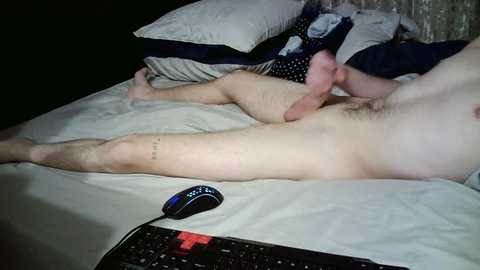 Video of a nude man with a flaccid penis lying on a bed, surrounded by pillows and a black keyboard.