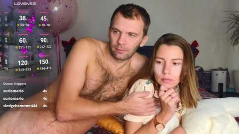 Video of a nude man and a woman in a bedroom, both smiling, with a digital overlay showing game stats.
