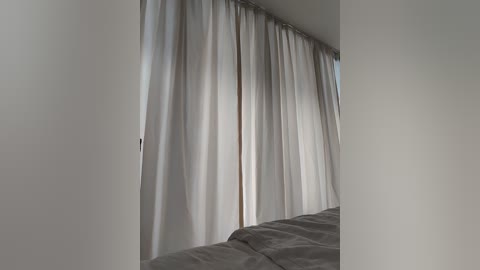 Media: Video of a minimalist bedroom corner featuring a bed with gray sheets partially visible. Tall, sheer white curtains with subtle horizontal lines are drawn, creating a soft, diffused light effect. The room has white walls, and the overall ambiance is calm and serene.