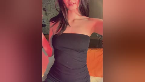 A video of a slender woman with long brown hair, wearing a strapless black dress, standing indoors. The background features a wall with a cartoon mural and a bed with orange sheets.