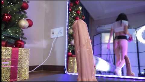Media: A video shows a woman in a mirrored room, wearing a white blindfold, black bikini, and high heels, with a large, erect, realistic-looking penis in front of her, Christmas tree, gifts, and festive lights in the background.