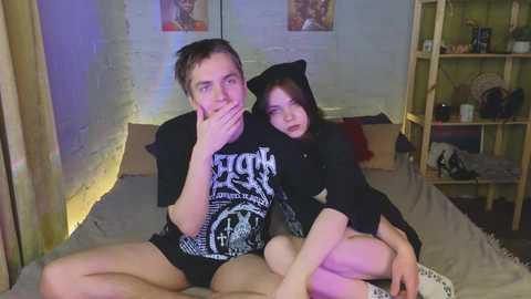 Media: Video of a young man and woman in a dimly lit, cluttered bedroom. He wears a black t-shirt, she a black hoodie with cat ears.