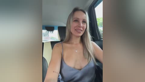 Media: A video of a smiling Caucasian woman with long blonde hair, wearing a low-cut blue camisole, sitting in a car with a blurred background.