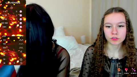 Media: Video of a young woman with long brown braids, wearing a sheer black top, talking into a microphone. Background shows a blurred bedroom with white bedding and a city skyline visible through a window.