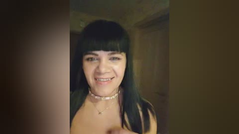 Video of a smiling woman with long, straight black hair and bangs, wearing a choker and a black top, indoors with wooden walls and a door in the background.