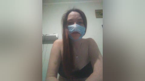 Video of a young woman with long brown hair, wearing a blue surgical mask, black strapless top, and a necklace, standing in a bathroom with a white sink and beige walls.