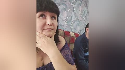 Video of a woman with straight black hair and bangs, wearing a purple lace dress, resting her chin on her hand, looking contemplative. Background features a patterned wall and a man in a blue jacket.