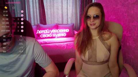 Video of a fair-skinned woman with long brown hair, wearing a beige halter top, sitting in a dimly lit room with pink lighting. A man in a grey shirt is partially visible in the background.