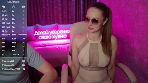 Video of a fair-skinned woman with long brown hair, wearing sunglasses and a sheer beige top, sitting on a chair with a pink neon sign in the background.