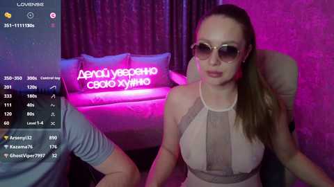 Video of a young, fair-skinned woman with long brown hair, wearing a sheer, nude-colored top, and large, round sunglasses, seated in a dimly lit room with pink and purple lighting.