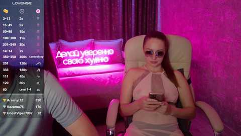 Video of a woman with fair skin, brown hair, and sunglasses, wearing a pink halter top and holding a phone, sitting in a beige armchair in a dimly-lit room with purple and pink lights.