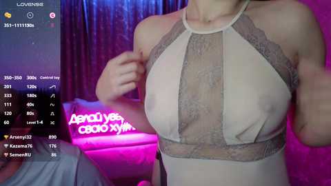 Video of a light-skinned woman in a sheer, beige lace top, showcasing medium-sized breasts, in a dimly lit, purple-lit room.