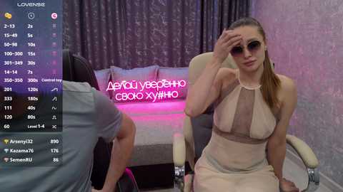 Video of a woman in a sheer beige dress with sunglasses, sitting on a beige chair, in a dimly lit room with purple curtains.