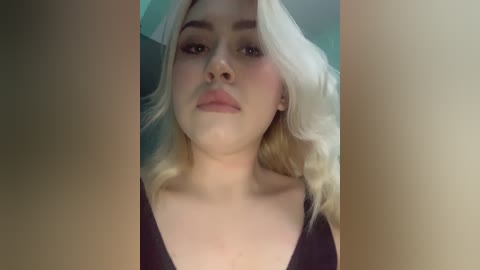 Media: Video of a young, fair-skinned woman with platinum blonde hair, wearing a black V-neck top, standing indoors with a blurred background.