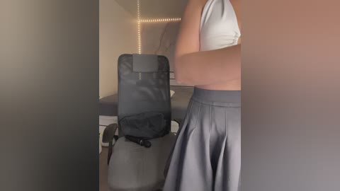 Media: Video of a woman in a white tank top and gray pleated skirt, sitting at a black office chair in a dimly lit room with beige walls and soft lighting.