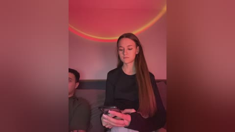 Media: Video of a young woman with long, straight brown hair, wearing a black top, seated next to a man with short black hair, both looking at a smartphone, in a dimly lit room with a red light.