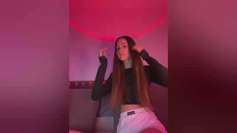 Media: Video of a young woman with long brown hair, wearing a black long-sleeve crop top and white shorts, sitting in a dimly lit room with pink and red lighting, adjusting her hair.