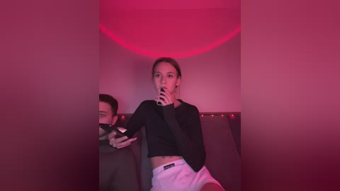 Media: Video of a young woman with dark hair, wearing a black crop top and white shorts, holding a smartphone, in a dimly lit, red-hued room.