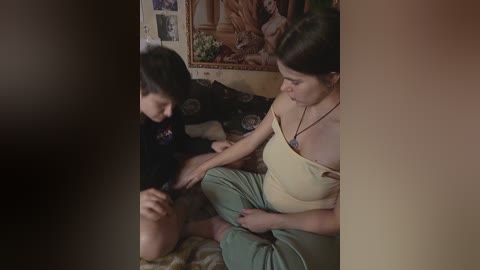 Video of a young woman with medium build, dark hair, wearing a beige tank top and green pants, assisting a young boy with a dark ponytail, dressed in a black hoodie, sitting on a couch in a dimly lit, cozy room with a framed painting on the wall.