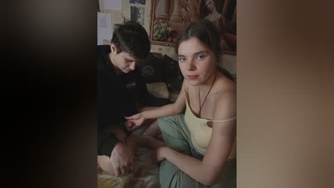 Video of a young woman with long brown hair, wearing a beige tank top and green pants, sitting next to a boy with curly hair in a dark room.