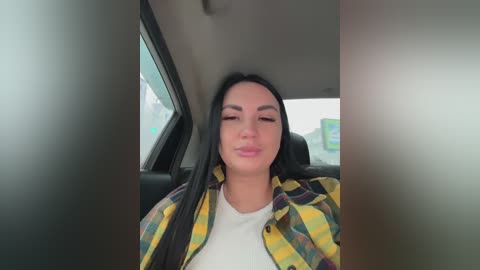 Video of a young woman with straight black hair, light skin, and a white top, sitting in a car, wearing a yellow and green plaid shirt, making a kissing face.