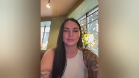 Video of a young woman with long black hair, wearing a white tank top, with tattoos on her arm, standing indoors near a window with green curtains.