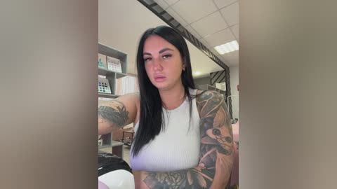 A video of a tattooed woman with long black hair, wearing a white tank top, sitting in a cluttered office with bookshelves and filing cabinets in the background.
