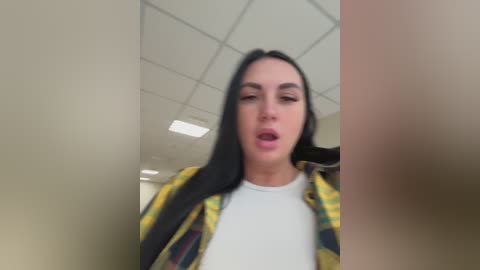 A video of a young woman with long black hair, wearing a white shirt under a yellow plaid shirt, taken from a low angle, showing her in a bathroom with tiled walls and a fluorescent ceiling.