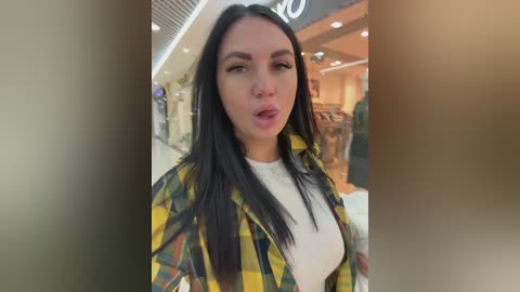 Video of a young woman with long black hair, wearing a yellow plaid shirt over a white top, standing in a brightly lit, modern shopping mall.