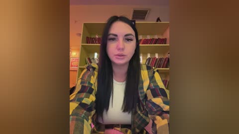 A video of a young woman with long black hair, wearing a yellow plaid shirt over a white top, sitting in a book-filled room with warm lighting.