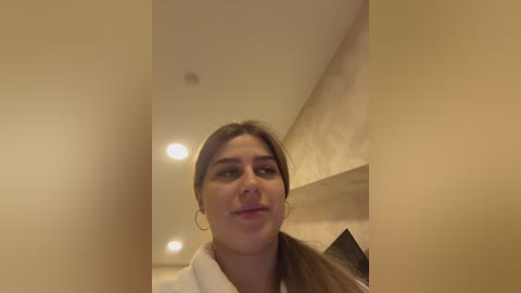 Media: A video of a young woman with brown hair, wearing a white robe, standing in a beige, dimly lit hallway with recessed ceiling lights.