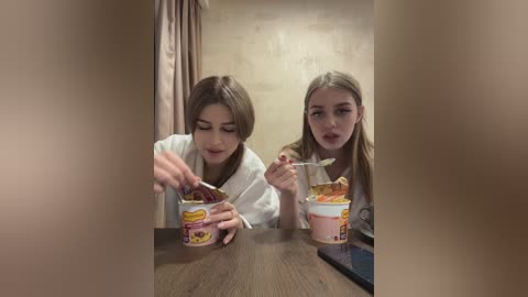 Media: Video of two young women with light skin, long hair, eating noodles from bowls in a dimly lit room with beige walls.