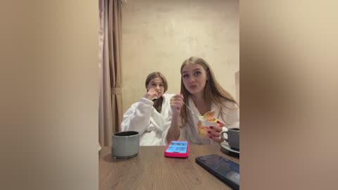 Media: Video of two young girls with long brown hair in white robes, eating ice cream and drinking coffee at a wooden table with a blue phone and mugs.