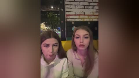 Media: Video of two young women, one with long brown hair, the other with light brown hair, wearing matching white sweaters, seated on a yellow couch in a dimly lit, brick-walled bar at night.