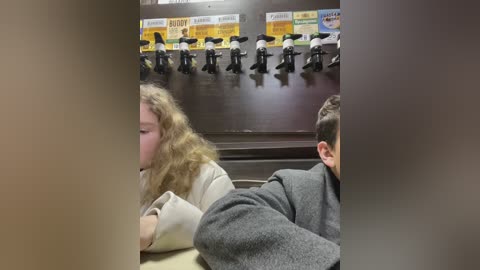 Media: Video of a dimly-lit room with two people in the foreground, one with curly blonde hair and the other with short brown hair, wearing gray sweaters. Above, a row of black shoes hangs on a wall.